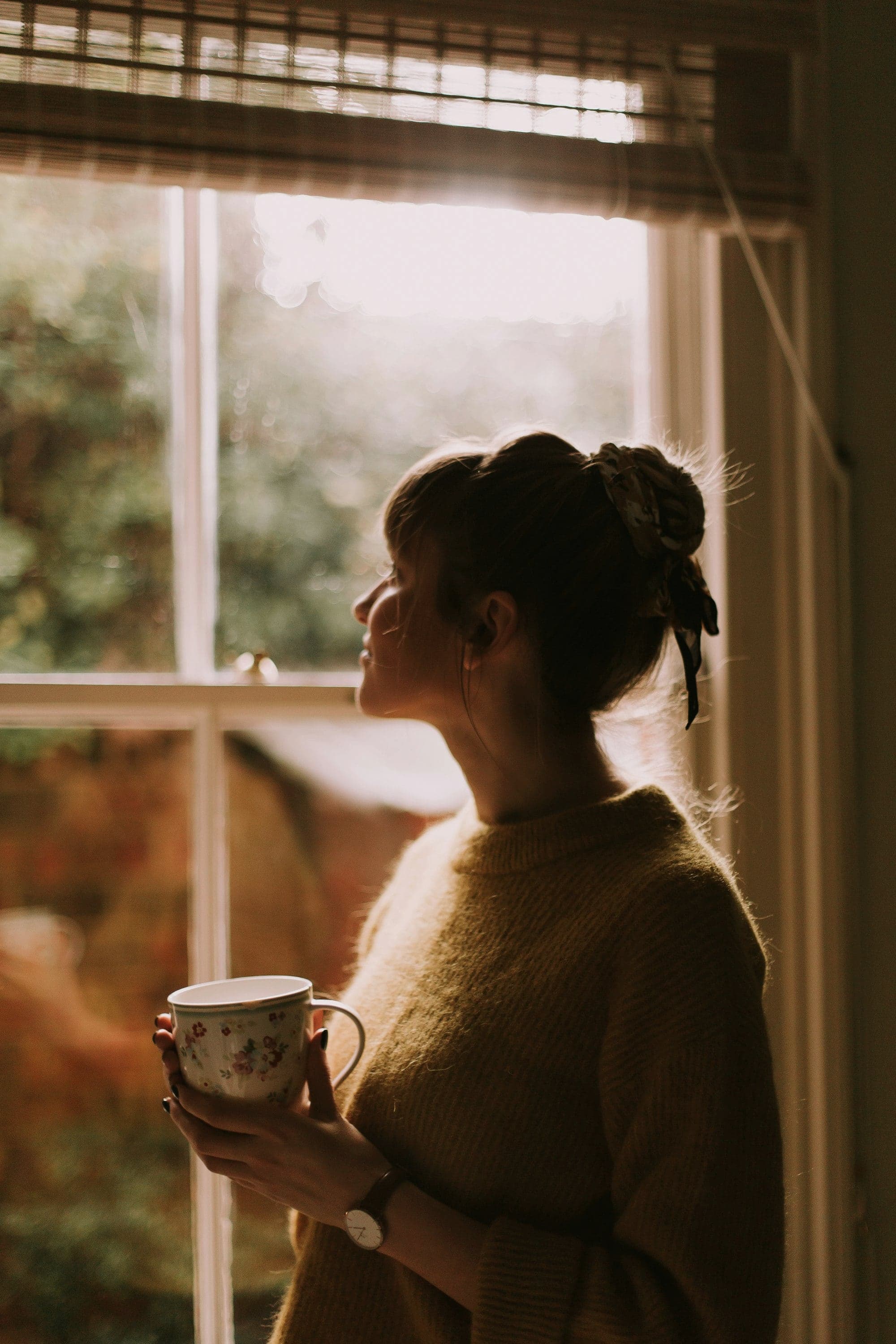 A woman holding a cup of tea, looking out a window in soft morning light